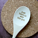 White spoon with 'just here to stir the pot' text on a cork coaster