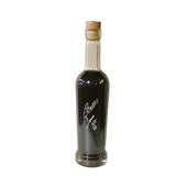 A clear glass bottle filled with a very dark brown liquid, sealed with a cork stopper, and featuring a white script logo on the front, isolated on a white background.